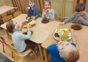 Dzieci siedzą wspólnie przy stole i degustują owoce: jabłka, banany, śliwki, brzoskwinie, mandarynki